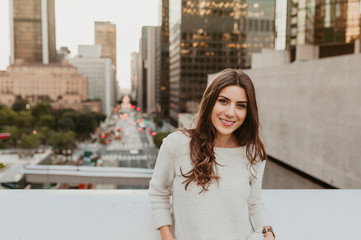 Beautiful young woman sitting on a bridge across the boulevard in urban scenery, downtown, at sunset, smiling at camera.