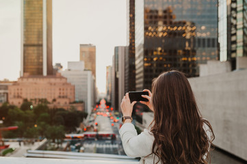 Beautiful young woman sitting on a bridge across the boulevard in urban scenery, downtown, at sunset, taking photos with smartphone.