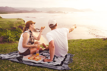 Cheerful couple spending nice time together while sitting on the beach and eating pizza