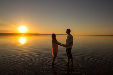 Young couple is holding hands in the water on summer beach. Sunset over the sea. . Two silhouettes against the sun. Calm and still surface of water. Romantic love story.
