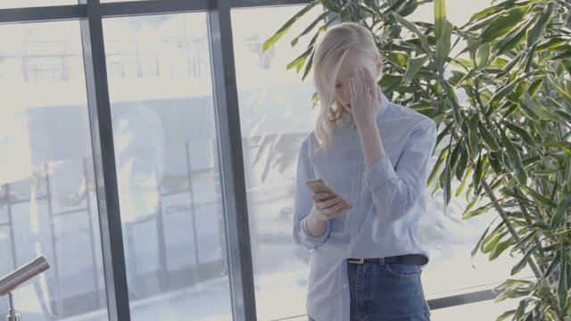 Portrait of female designer is typing messeges on her smartphone in bright hall, slow motion. Young lady with blond hair, in blue shirt and jeans is standind near the big window and troical plant and