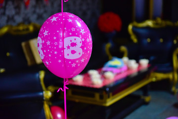A pink and magenta balloon inflated with helium and the number eight on it for a birthday party. In the blurred background, black and gold armchairs and pop corn above the table