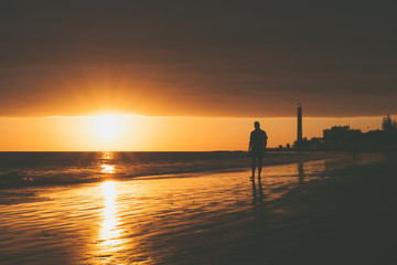 Walker on seashore with lighthouse during sunset in Maspalomas, Gran Canaria Island.