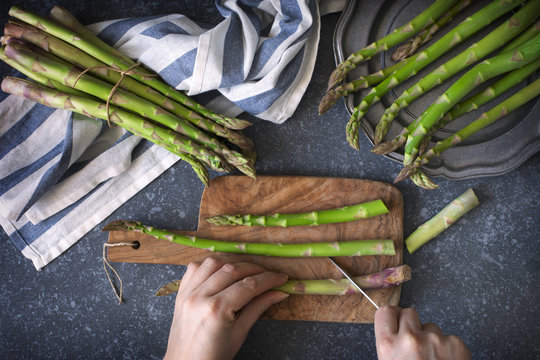 Fresh  raw asparagus on stone background. Women female hands cut asparagus on wooden cooking board. Top view