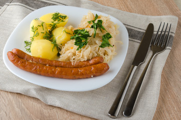 Roasted sausages with steamed cabbage (sauerkraut) and boiled potatoes in white plate.