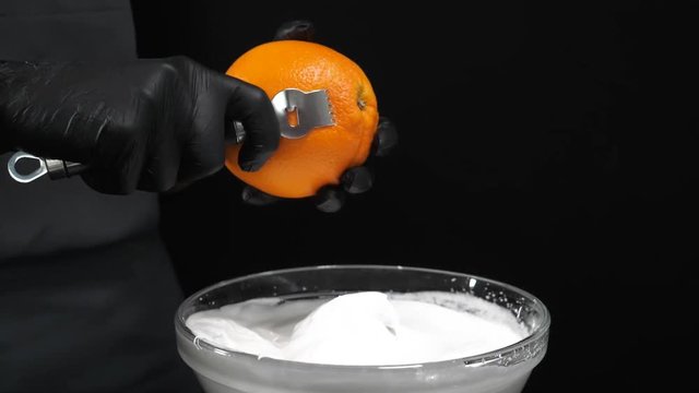 Slow motion of citrus zester grating peeling orange peel, oils spraying against dark background
