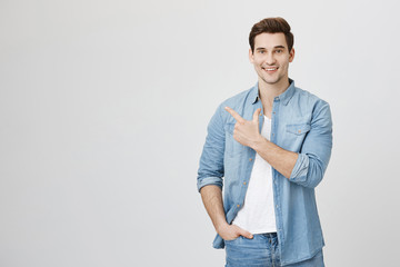 Close-up portrait of healthy handsome caucasian guy with brown hair in denim clothes pointing with one hand and holding another in pocket, being happy and smiling broadly over white background