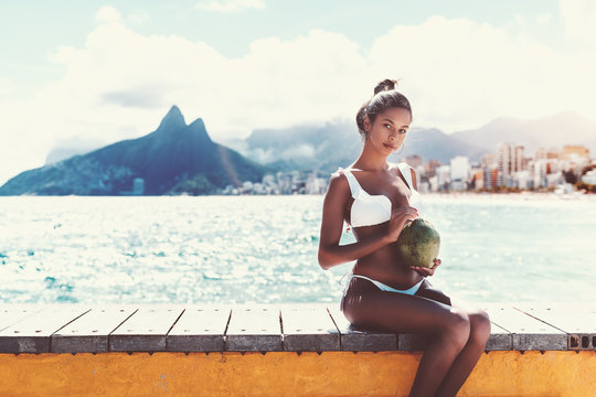 Charming young Brazilian girl is sitting on wooden bench next to beach and holding green coconut for drinking with cityscape of Rio de Janeiro and the Two Brothers hills in blurry background