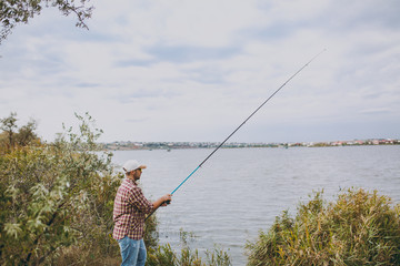 Side view Young unshaven man with a fishing rod in checkered shirt, cap and sunglasses casts a fishing pole on lake from shore near shrubs and reeds. Lifestyle, recreation, fisherman leisure concept