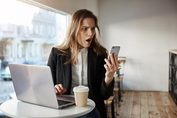 Oh my, I am late for work. Portrait of shocked and surprised stylish woman in cafe, sitting near laptop and drinking coffee, looking at smartphone screen with amazed and afraid expression