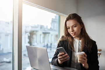 Surrounded with gadgets. Indoor portrait of attractive caucasian businesswoman sitting in cafe near window, drinking coffee and messaging via smartphone, working with laptop during break