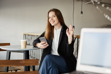 Handsome guy with laptop tries to become friends with attractive woman. Portrait of cute european girl sitting in cafe, using wifi with smartphone and drinking coffee, waving at camera while smiling