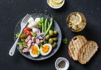Fototapeta na wymiar Aperitive table - plate of canned tuna, green beans, mozzarella cheese, tomatoes, boiled egg, olives, grilled bread and whiskey glasses on dark background, top view