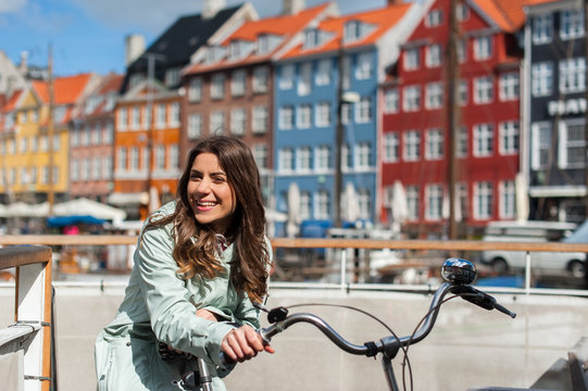 Young happy woman on bicycle at the Nyhavn harbor pier in european city Copenhagen, Denmark, looking at camera and smiling. Visiting Scandinavia, famous European place.