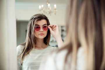 Naklejka premium Attractive woman on hunt for new trendy things. Indoor portrait of stylish young caucasian woman in optician store trying on fashionable sunglasses while standing near mirror, picking from variants