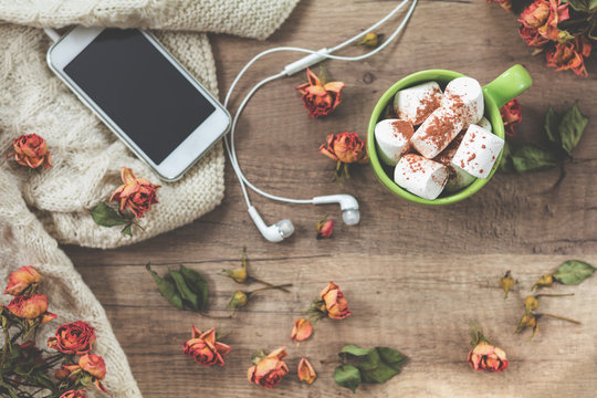 Cup of coffee with marshmallow, white knitting wool, dried roses flowers, mobile phone and headphones