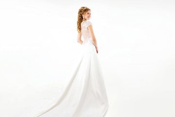 Fototapeta na wymiar young blonde girl in a white wedding dress and jewelry in hairstyle with big curls standing from the back like princess on a white wall background 