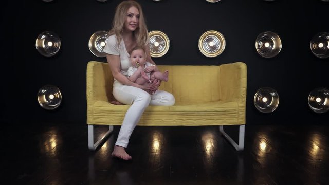 Lovely mother in white, her little baby and a dog come and amicably sit down on the yellow sofa