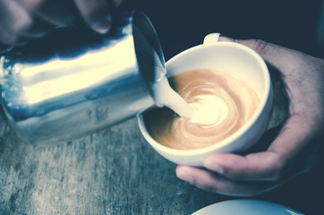 Closeup image of male barista holding and pouring milk for prepare cup of coffee, latte art, vintage color tone, coffee preparation and service concept, lifestyle