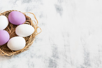 White and purple Easter eggs lying on a tray made of twine, under them hay. White wooden vintage...