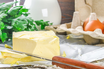 A bar of butter is cut into pieces on a wooden board with a knife, surrounded by milk, eggs and parsley on a brown table. Ingredients for cooking.