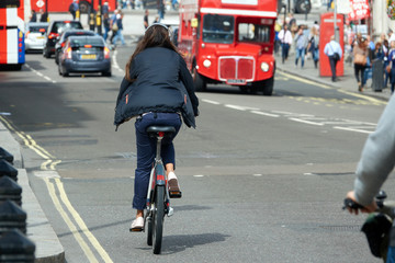 A girl in white headphones rides a bicycle on the streets of London on the background of a red bus