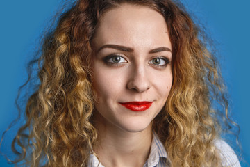 Close up portrait of happy friendly looking young lady with voluminous wavy hair and red lips relaxing indoors against blue wall background, having joyful positive expression on her pretty face