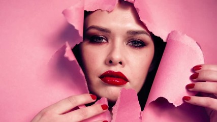 Face beautiful girl with bright make-up, with pouting lips,red lipstick,fingers with red nails, appears in torn opening in pink paper background.Creative advertising women's cosmetics. Girl peek hole.