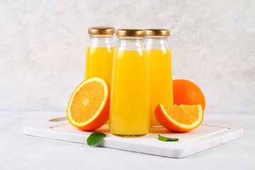 Glass bottles with fresh orange juice with orange slices and yellow tubes on a light gray table.