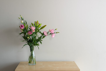 Pink lisianthus flowers and green foliage in glass vase on wooden shelf against beige wall