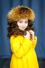 A photo of a beautiful little girl with blue eyes and brunette curly hair in studio wearing a fur hat and yellow coat