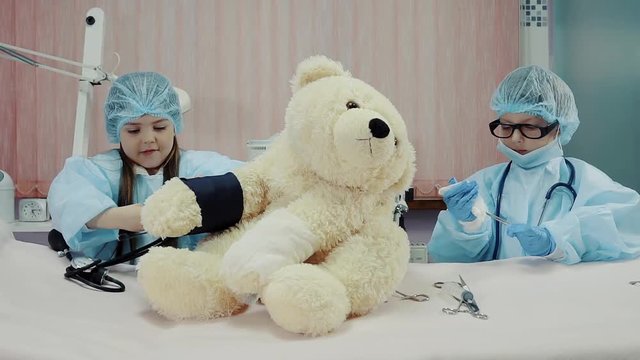Cute girl is measuring pressure of a big teddy bear. Children is playing in a physicians, little boy is preparing tools for imaginary operating