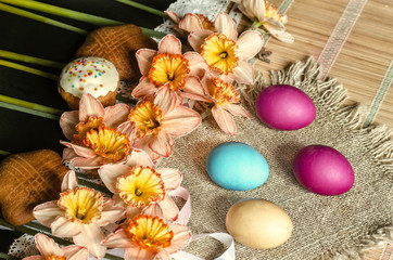 Fototapeta na wymiar Large pink daffodils with yellow fringe in the middle and biscuits on burlap with painted Easter eggs lie on straw carpet