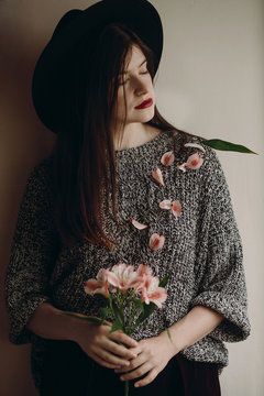 stylish hipster girl in hat holding pink flowers in room. boho woman holding beautiful alstroemeria in hands in spring morning and petals on body. creative sensual female portrait