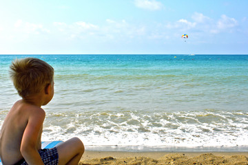 little boy sits on the beach and looks at the sea and parachute in the sky