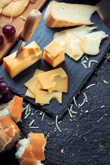 Black slate board with various cheese, decorated with grapes, bread and honey