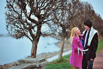 Love story of young beautiful couple in Peterhof, Saint-Petersburg, Russia at autumn. Blonde girl in pink coat and blue dress and man or groom in black jacket and jeans. Embrace kiss with fall in love