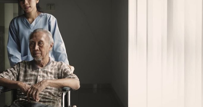 Slow motion of elderly man sitting on wheelchair and pushed by nurse near the window in the hospital. Shot in 4k resolution
