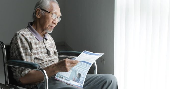 Senior man sitting on wheelchair while reading a newsletter and wearing glasses near the window at home. Shot in 4k resolution