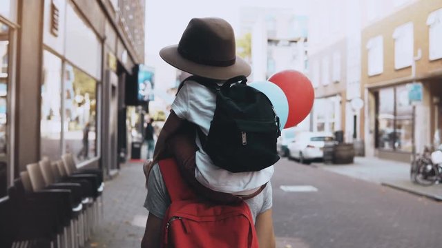 Boy child on mom's shoulders, two air balloons. Mother and son in hat with backpack walk together along the street. 4K.