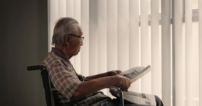 Lonely old man with grizzled hair, sitting on wheelchair while wearing glasses and reading a newspaper near the window. Shot in 4k resolution