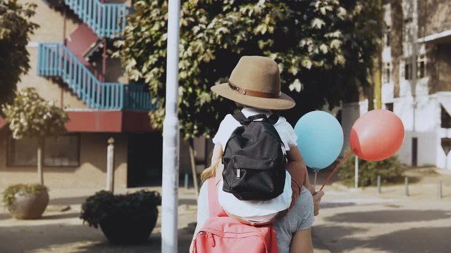 Mother carries son on shoulders among houses. Woman walking with a kid in hat and two air balloons. Lifestyle 4K.