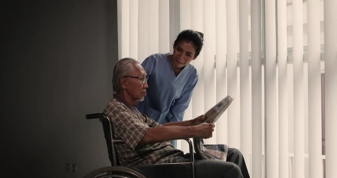 Old man sitting on wheelchair and pushed by his assistance or nurse after reading a newspaper near the window. Shot in 4k resolution