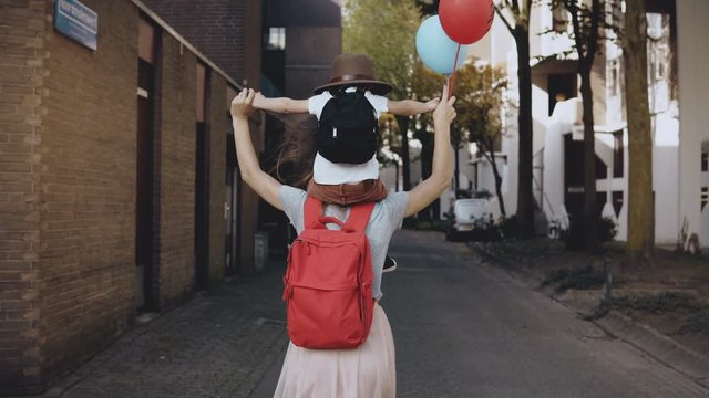 Mom with son on shoulders walk holding hands. Happy woman carries a kid in hat and two air balloons. Lifestyle 4K.
