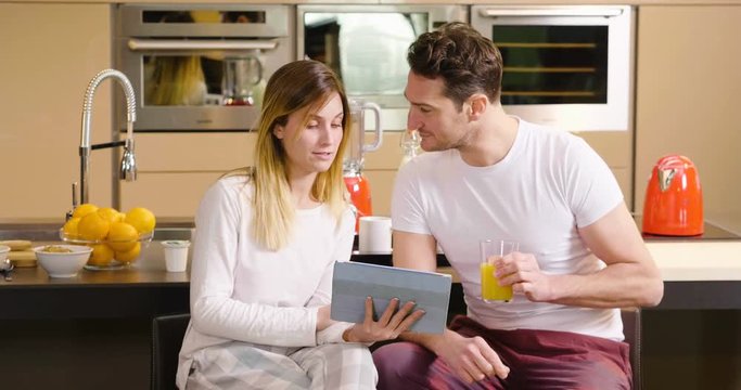 A couple in the kitchen looks at the tablet with the souvenir photos of their holidays or of the past times while having breakfast and smiling happily. Concept of: family, technology, memories