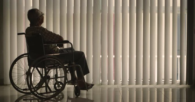 Silhouette of elderly man looking at the window with curtain while sitting on wheelchair and asleep. Shot in 4k resolution