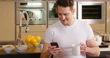 A man in the kitchen while having breakfast and sipping tea or milk in the cup sends a message or calls with the phone and smiles. Concept of: social network, message, technology.