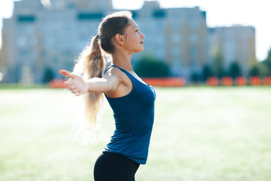 Young fitness woman in a blue shirt and leggings smile spread her hands to the sky for success in waiting the sun shines in her face, in stadium on the background of houses.