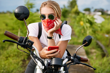 Fototapeta premium Fashionable female driver dressed casually, reads bikers blog on mobile phone, sits on motorbike, freshes fresh air outdoor, looks thoughtfully into distance. People, lifestyle and technology