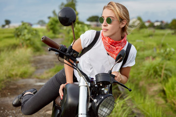 Fototapeta na wymiar Fashionable young female biker makes stop on long way, stands up from motorbike, wears shades and casual comfortable clothes, has confident look away, rides in countryside. Lifestyle concept